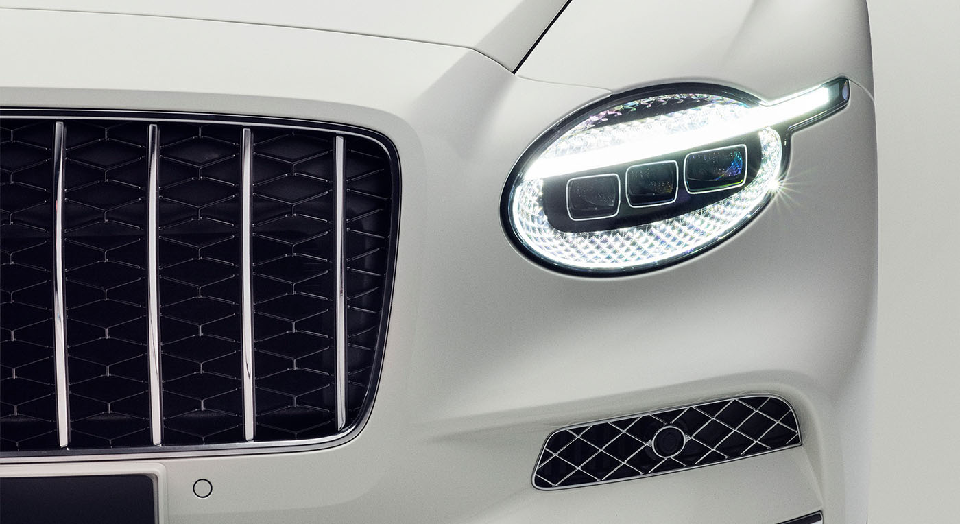 Close-up of a sleek white car's front, showcasing a modern, oval LED headlight with a grid-like pattern and a black grille with vertical chrome bars.